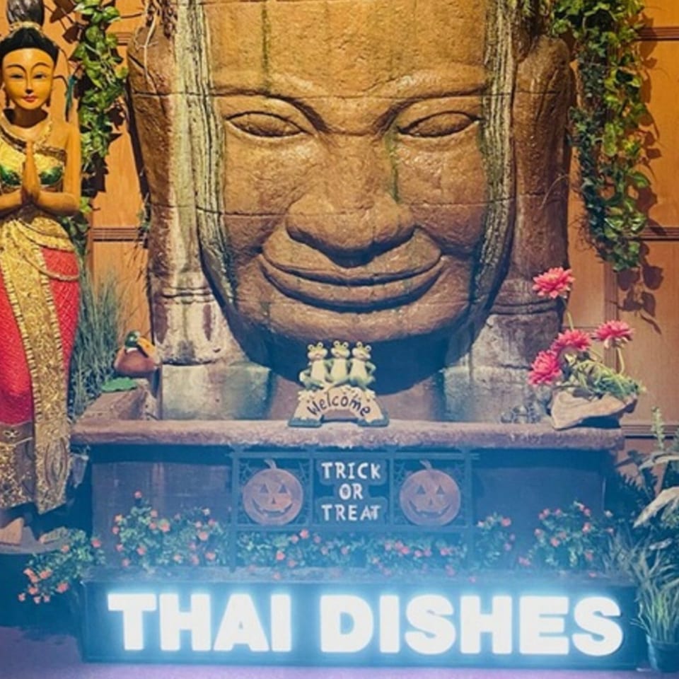 A large stone Buddha head with a Thai statue, plants, and a glowing "THAI DISHES" sign.