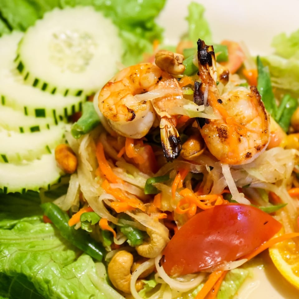Close-up of a Thai salad with grilled shrimp, shredded papaya, cashews, and cucumber slices.