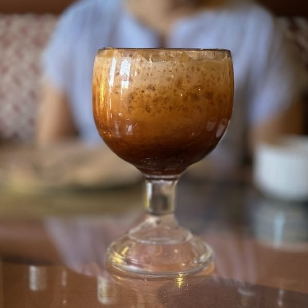 A glass goblet filled with a dark brown, frothy beverage sits on a table.
