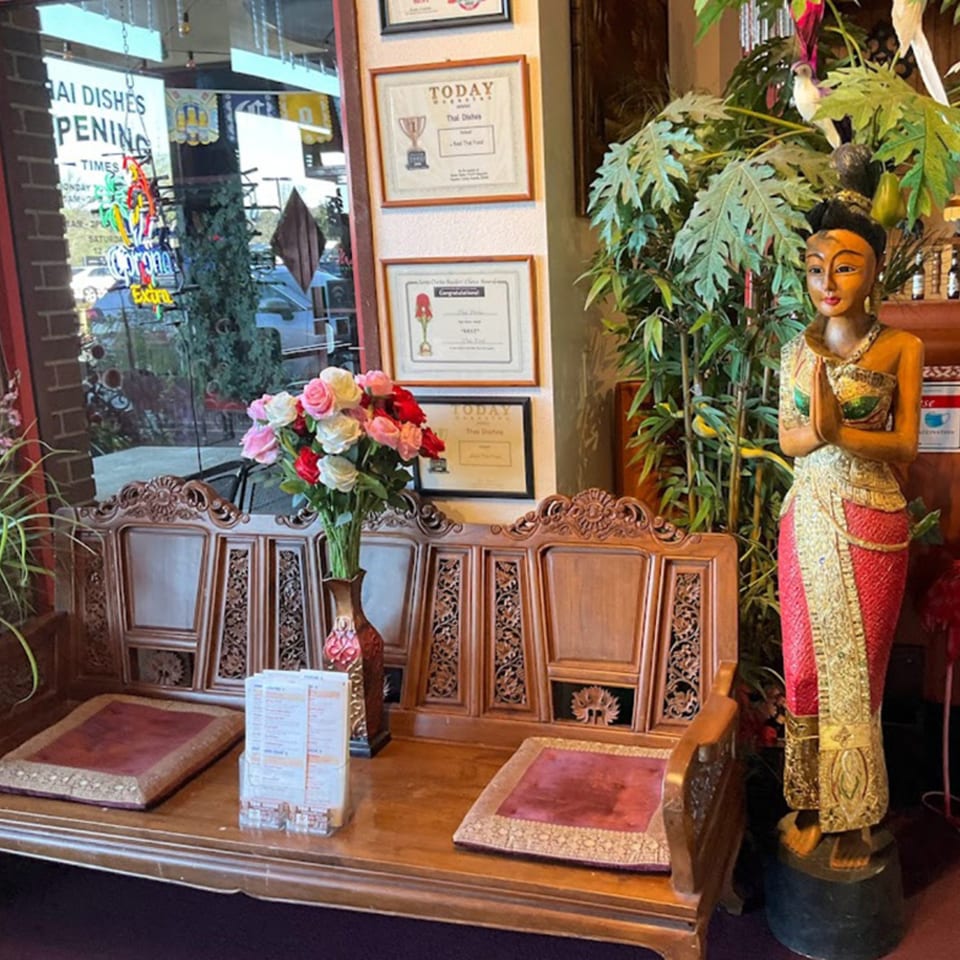 Ornate wooden bench with red cushions, a vase of roses, and a Thai statue in a restaurant.