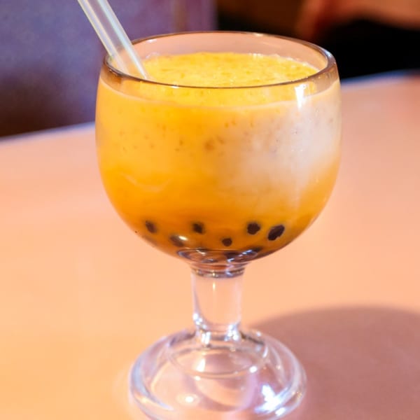 A Thai mango boba drink with a straw in a stemmed glass on a peach-colored table.