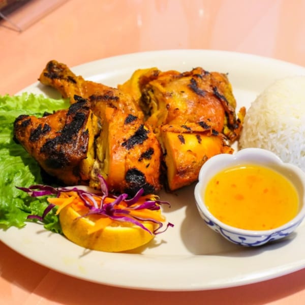 Grilled Thai chicken with rice, lettuce, lemon, and a side of dipping sauce on a white plate.